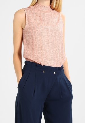 Femme portant un chemisier sans manches à fleurs rose rentré dans un pantalon large taille haute bleu foncé, les mains dans les poches.
