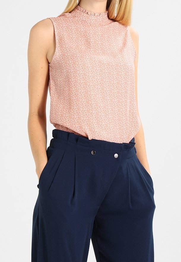 Femme portant un chemisier sans manches à fleurs rose rentré dans un pantalon large taille haute bleu foncé, les mains dans les poches.