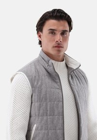 Graue, gesteppte Weste mit hohem Kragen und Reißverschluss, getragen über einem weißen, strukturierten Strickpullover. Verfügt über Seitentaschen und kontrastierende Kanten.