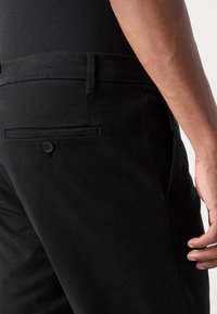 Gros plan sur une personne portant un pantalon noir avec une poche arrière boutonnée et une chemise noire rentrée, main reposant près de la cuisse.