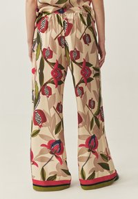Vrouw in beige wijde pantalons met rode en groene bloemmotieven, staande met ontspannen handen langs de zij, met de rug naar de kijker gekeerd.