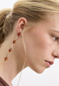 Goldfarbene, hängende Ohrringe mit geometrischen braunen Harzverbindungen in Diamantenform erzeugen ein längliches Design, das einen Kontrast zur Haut bildet.