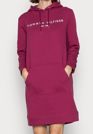 Kobieta w długim, kapturowym, ciemnoróżowym sukience typu sweatshirt Tommy Hilfiger z przednią kieszenią, ręce częściowo w kieszeniach.
