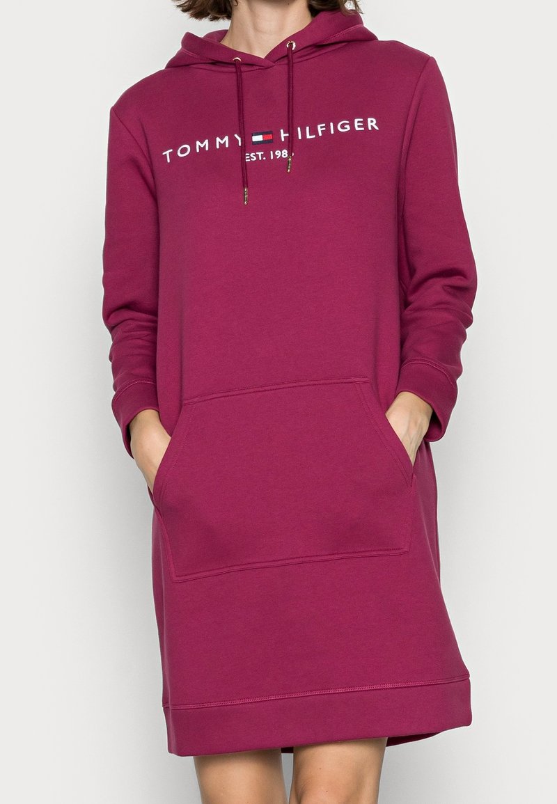 Femme portant une robe sweat à capuche manches longues Tommy Hilfiger rose foncé avec poche avant, mains partiellement dans les poches.