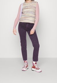 Gilet lavorato a maglia in filato pastello multicolore sopra una lunga maglia rosa, abbinato a jeans viola e sneakers alte con motivi e dettagli rosa.