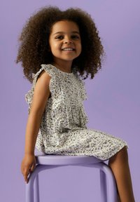 Valkoinen mekko, jossa on pieniä vihreitä ja purppuran värisiä kukkakuvioita, röyhelöhihoilla, jota tyttö pukee purple tuolilla, vaalean purppuran taustan edessä.