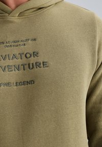 Zeleno-olivová mikina s kapucí vyrobená z měkké tkaniny s volným střihem. Na hrudi je vyražený text: "AVIATOR ADVENTURE" a "TYPE No. C46".