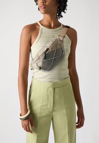 Beige crossbody bag made of soft fabric with a mesh pocket, featuring a zipper closure and elastic accents, worn over a striped tank top.