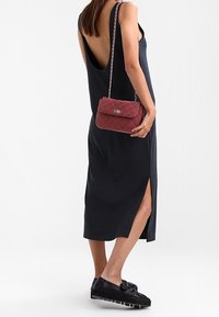 Donna in un vestito midi senza maniche nero con schiena scoperta e spacco laterale, che tiene una borsa a tracolla trapuntata bordeaux e indossa mocassini neri con plateau.