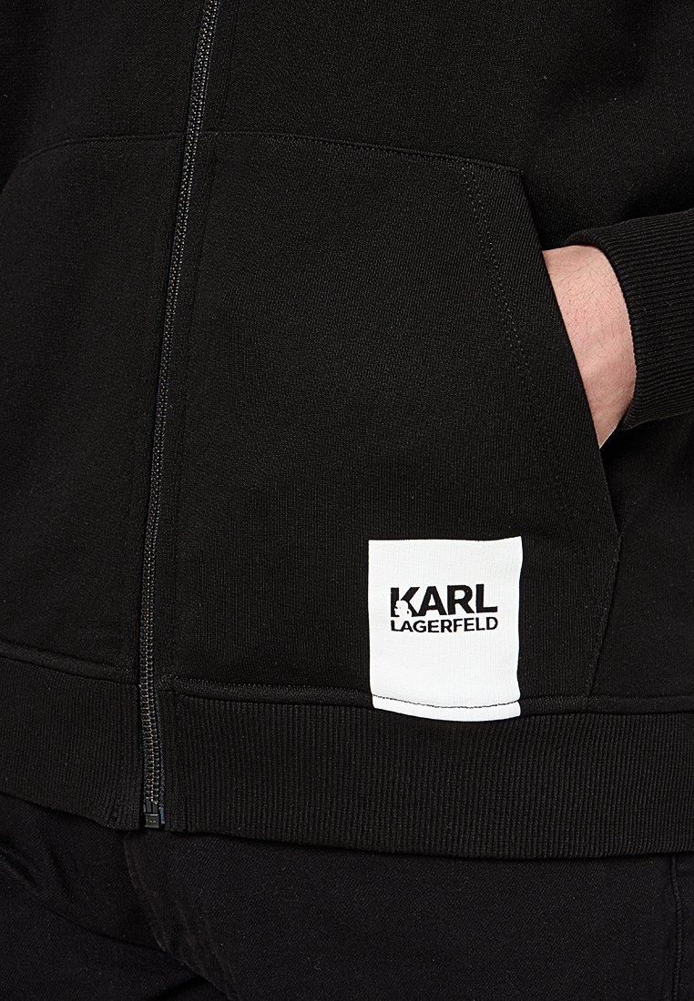 Sudadera con cremallera negra, con puños y dobladillo acanalados. Presenta un parche con logo blanco en el bolsillo delantero. El material parece suave y texturizado.