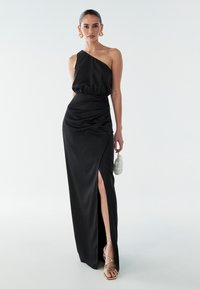Schwarzes Satin-Ein-Schulter-Kleid mit geraffter Taille und einem hohen Schlitz bis zum Oberschenkel. Kombiniert mit goldenen Sandalen mit Riemen und einer kleinen grünen Tasche.