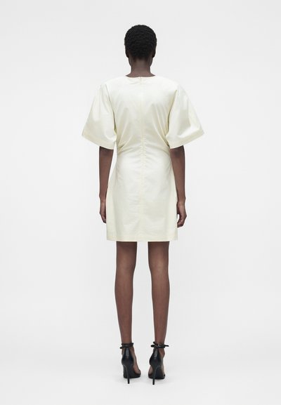 Άτομο με κοντά σγουρά μαλλιά που φοράει μια κρεμ κοντή φόρεμα με φουσκωτά μανίκια και μαύρα ψηλοτάκουνες γόβες με λουράκι στον αστράγαλο, βλέποντας από πίσω.