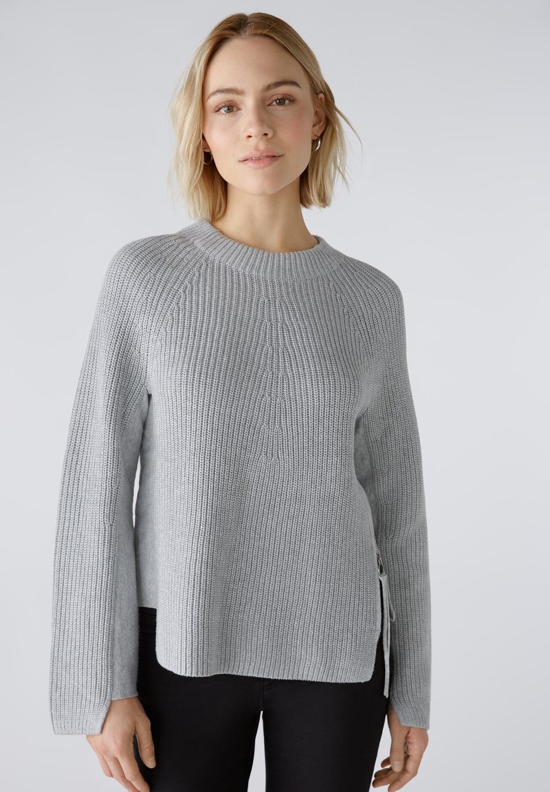 Pull en maille gris clair avec une texture côtelée, décolleté rond et fentes sur les côtés. Présente des manches longues et une coupe décontractée.