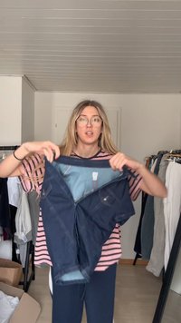 Young woman with glasses holding up dark blue denim shorts in a room with clothes racks and an open cardboard box.