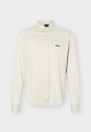 Biała, długorękawowa koszula zapinana na guziki z kołnierzykiem, czarne logo "BOSS" na lewej piersi oraz mankiety zapinane na guziki, prezentowana na jednolitym tle.