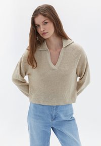 Krämfärgad stickad tröja med V-ringning och vikbar krage. Lätt lös och kort passform med långa ärmar och ett texturerat mönster. Ljusblå jeans.