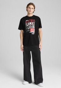 Sort T-shirt med et farverigt trykt design med teksten "FORZA MILAN". Kombinationen inkluderer vide sorte jeans og hvide sneakers.