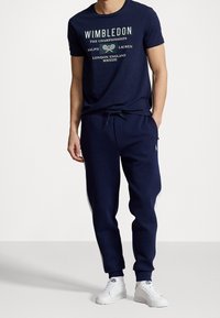 Muž oblečený v námořnickém tričku Wimbledon, s odpovídajícími joggingovými kalhotami a bílými teniskami, stoj í na jednobarevném pozadí, s jednou rukou v kapse.