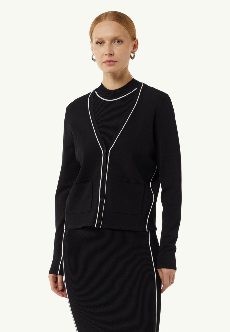 Schwarze Strickjacke mit weißem Besatz, V-Ausschnitt, zwei vorderen Taschen und Knopfverschluss. Rippstruktur mit taillierter Passform.