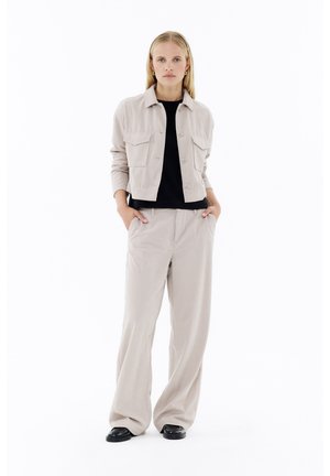 Beige Cordjacke aus Kord mit zwei Fronttaschen, getragen über einem schwarzen Hemd. Weite beige Cordhosen kombiniert mit schwarzen Schuhen.