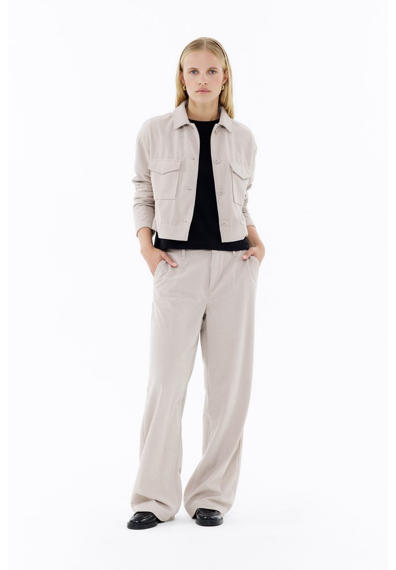 Beige Cordjacke aus Kord mit zwei Fronttaschen, getragen über einem schwarzen Hemd. Weite beige Cordhosen kombiniert mit schwarzen Schuhen.