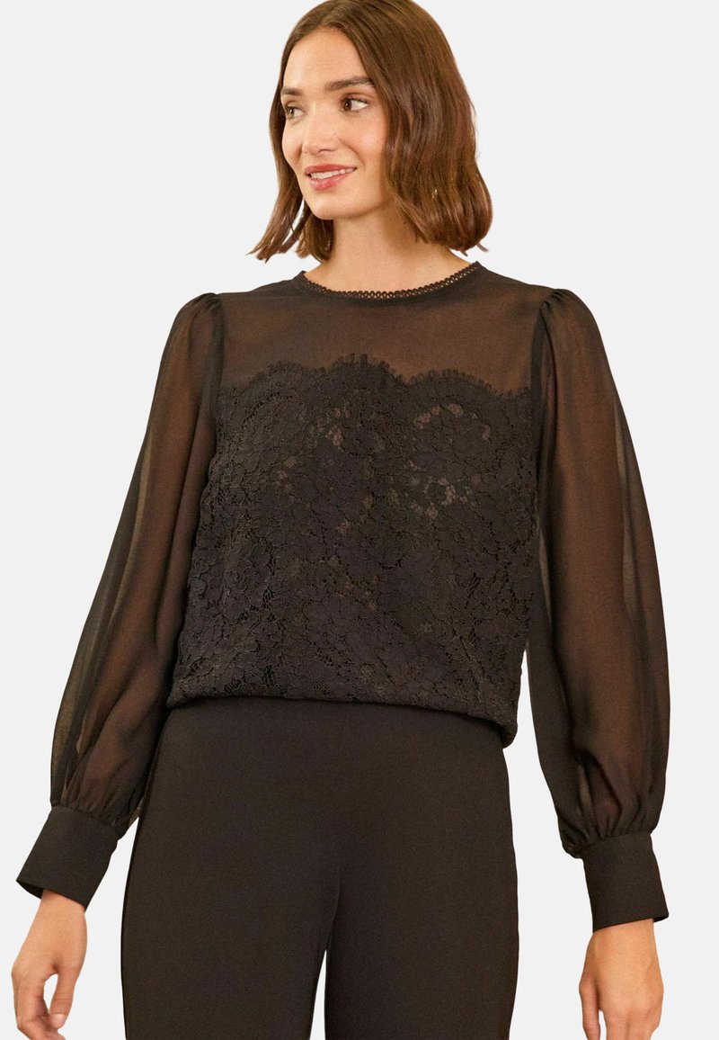 Blusa nera con maniche lunghe trasparenti, dettaglio in pizzo testurizzato sul capo, e scollatura rotonda con una sottile bordatura. Tessuto liscio sotto la vita.
