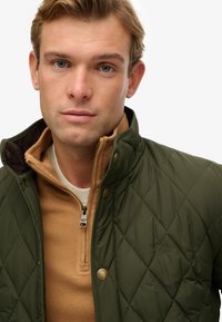 Joven con cabello rubio que lleva una chaqueta acolchada verde sobre un suéter beige con cremallera, contra un fondo blanco liso.
