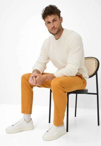 Crème textuur gebreide trui, oranje chinos en witte sneakers. Zittend op een zwarte stoel met een geweven leuning. Eenvoudig en modern ontwerp.