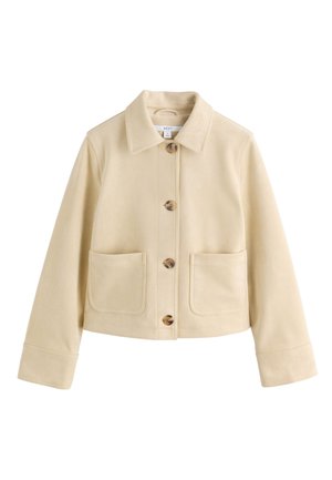Frau mit glatten schwarzen Haaren, die eine beige Jacke und einen dunkelbraunen Rock trägt, von hinten vor einem schlichten hellen Hintergrund dargestellt.