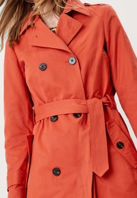 Červený trenčkot vyrobený z hladkého materiálu, s dvojitým zapínáním vpředu na černé knoflíky, pasem podtrženým páskem a bočními kapsami.