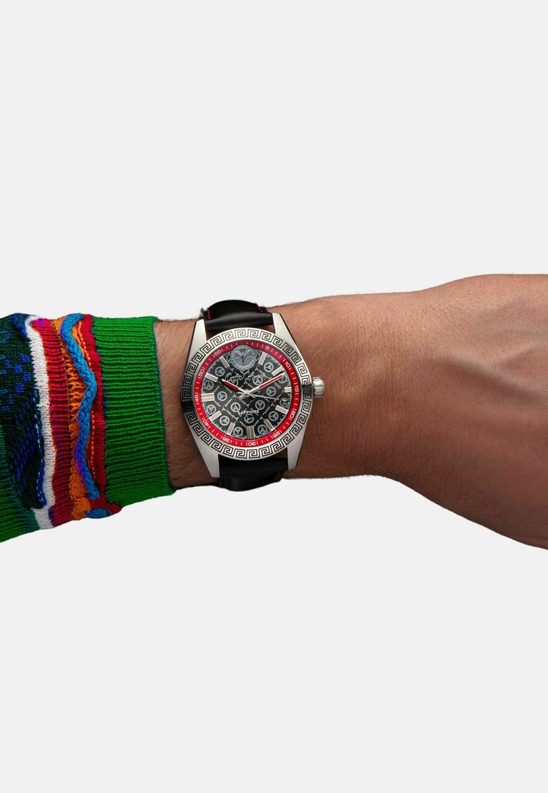 Silberne Uhr mit schwarzem Gesicht im Schädelmuster, roten Akzenten und griechischem Schlüsselrand. Schwarzes Armband mit roter Naht. Am Handgelenk in einem bunten Pullover getragen.