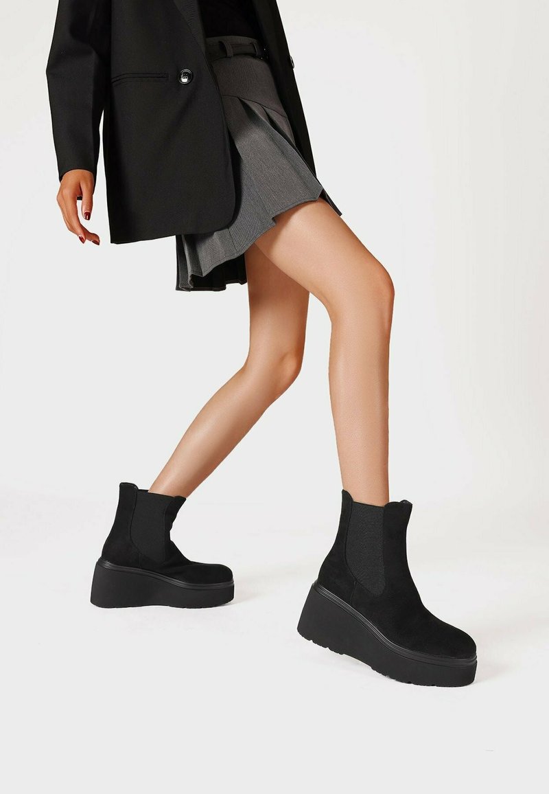 Stivali neri alla caviglia in suede con pannelli elastici e una suola platform spessa. Ambientati su uno sfondo neutro, abbinati a una gonna a pieghe.