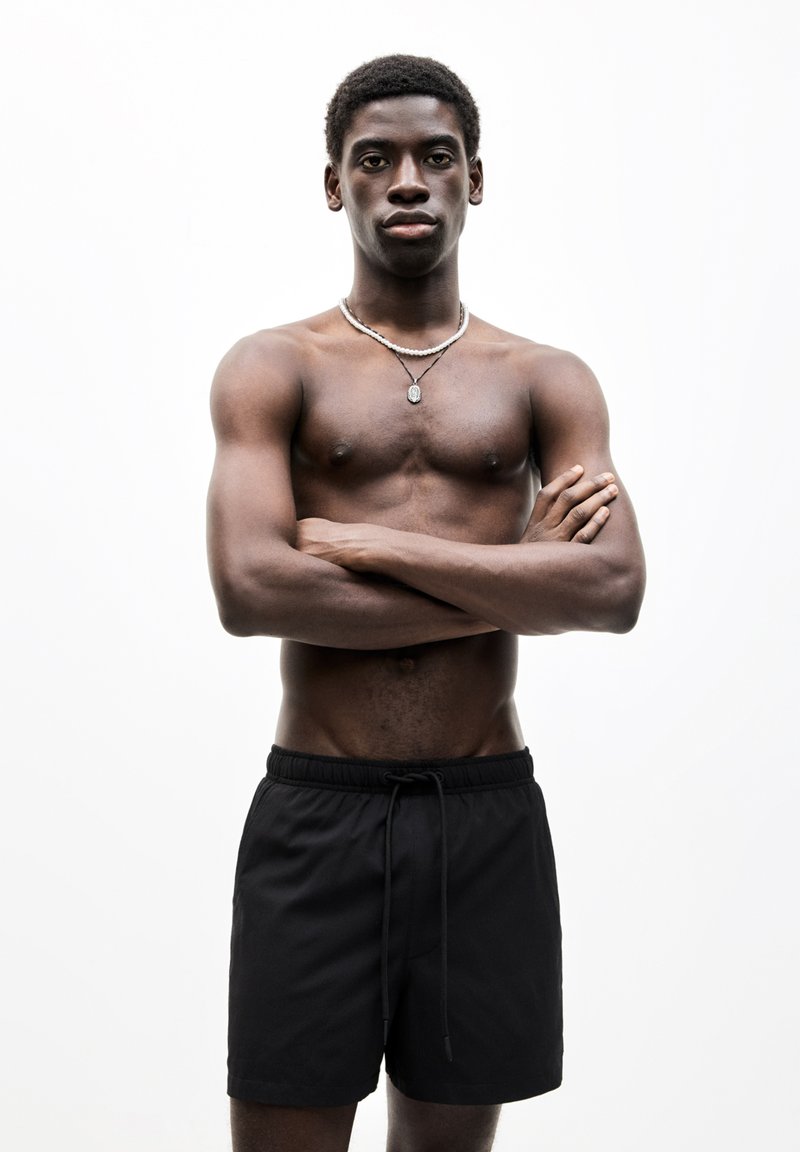 Schwarze Schwimmshorts mit elastischem Bund und Kordelzug. Das Model hat einen nackten Oberkörper, kurze, lockige Haare und trägt eine Halskette.