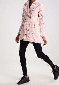 Svetle růžový trenčkot s páskem v pase, se zipovými kapsami a květinovou výšivkou na rukávech; spárovaný s černými legínami a teniskami.