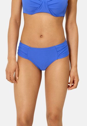 Mujer con bikini azul, con la parte inferior y superior a juego, sobre un fondo claro y sencillo, con las manos relajadas a los lados.