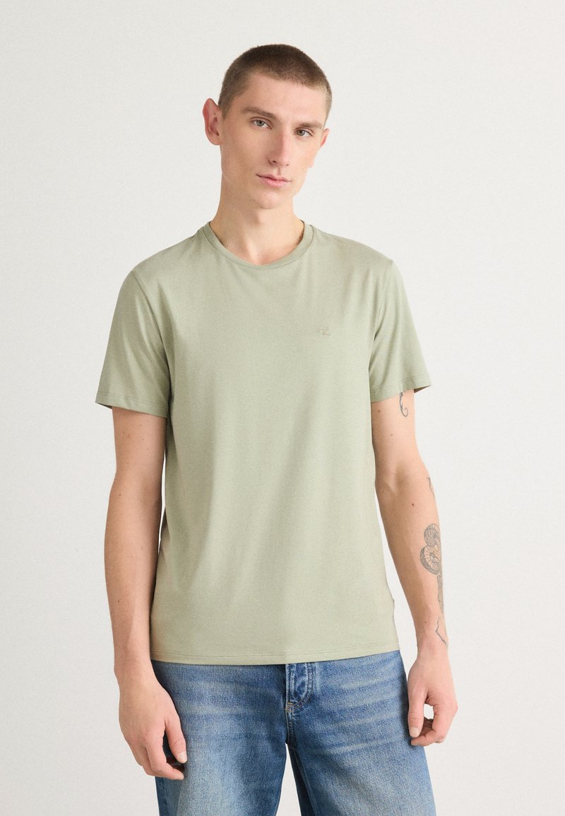 T-shirt vert clair à manches courtes en tissu doux, avec un col rond et un accent logo subtil sur la poitrine, porté avec un jeans bleu.
