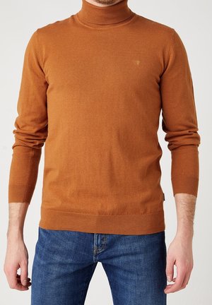 Hombre con un suéter de cuello alto ajustado de color naranja quemado y unos vaqueros azules, de pie con los brazos relajados a los lados frente a un fondo liso.