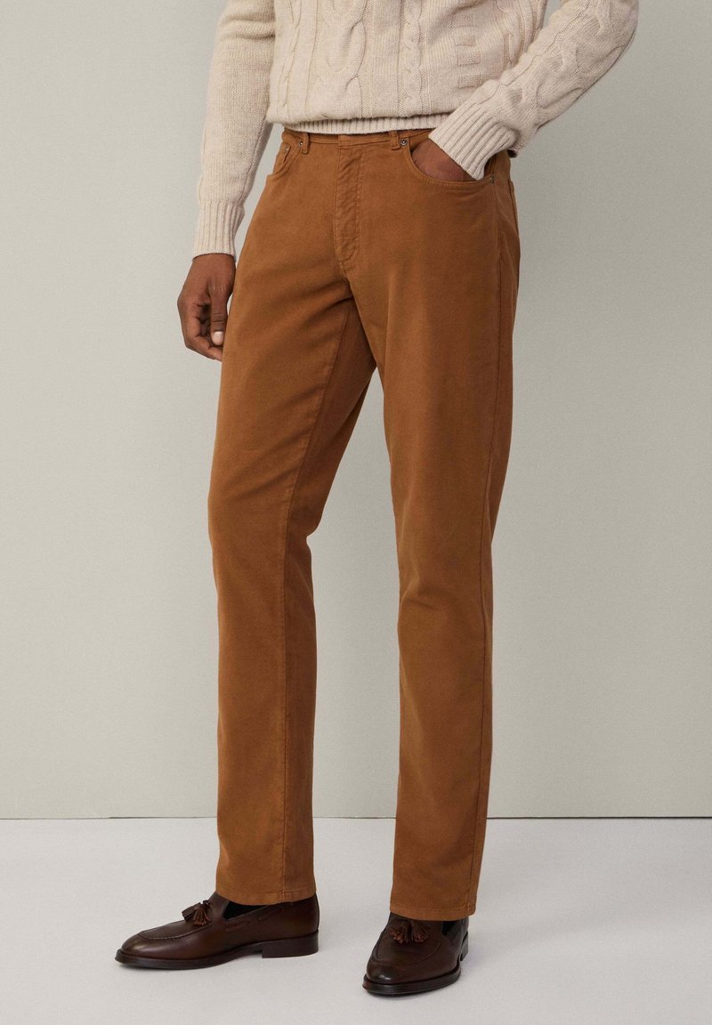 Smeđe pamučne chinos hlače slim fit kroja, s bočnim džepovima i glatkom teksturom. U kombinaciji sa tamnosmešnim mokasinkama.
