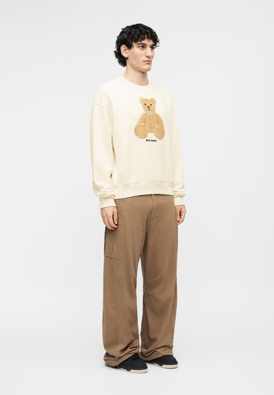 Νέος άνδρας στέκεται φορώντας μπεζ φούτερ με γραφική παράσταση αρκουδάκι και καφέ παντελόνι φαρδιάς γραμμής με μαύρα αθλητικά παπούτσια σε απλό φόντο.