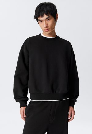 Young man wearing a loose black sweatshirt and matching pants, standing against a plain light gray background with a serious expression.