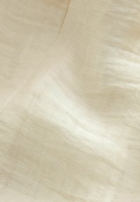 Tissu beige texturé avec une légère brillance et un motif de tissage visible, délicatement drapé pour créer des plis et des ombres douces.