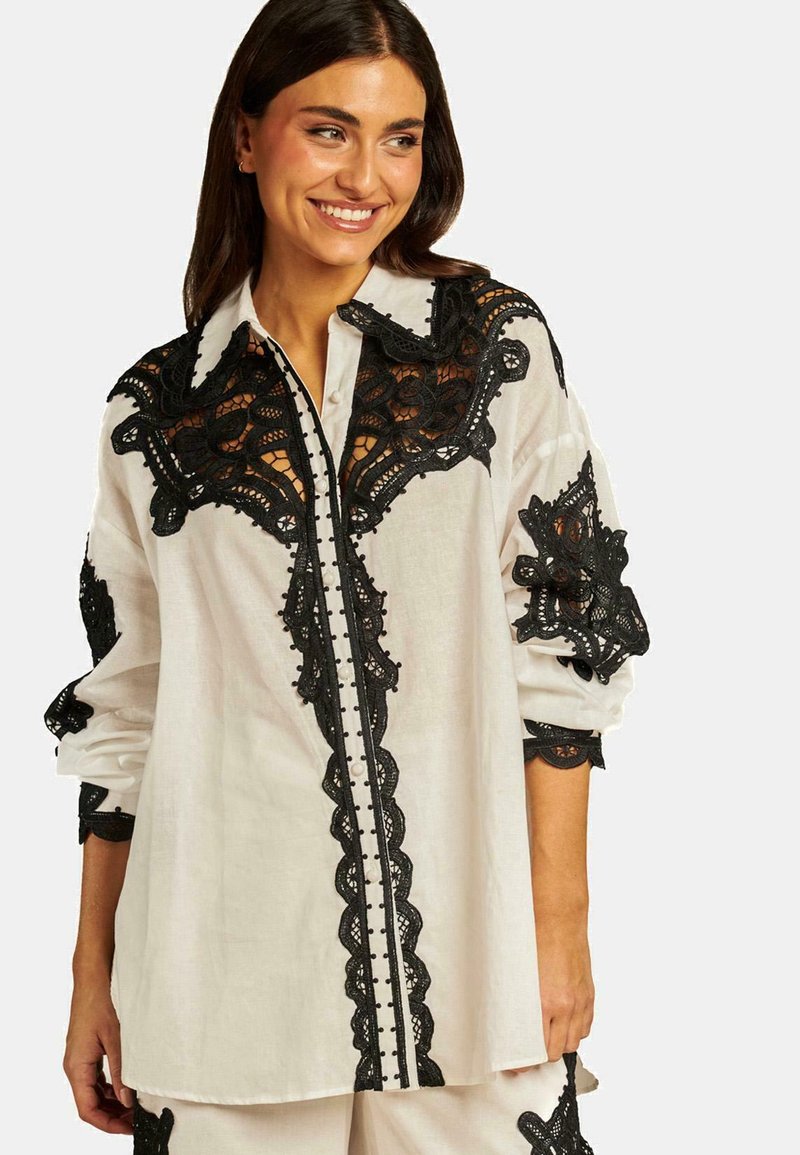 Camicia bianca con dettagli in pizzo nero intricati sul colletto e sulle maniche; presenta chiusure a bottoni e una vestibilità morbida.