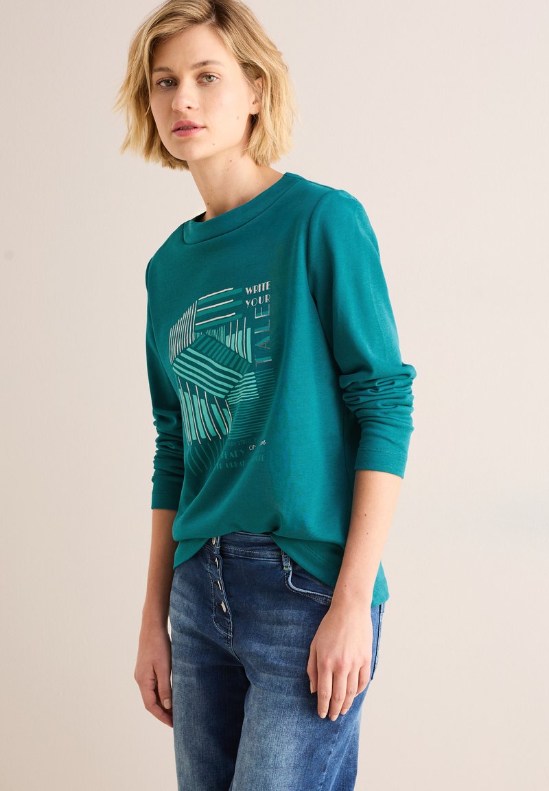 Teal langekleurig shirt met geometrische grafische print, ronde halslijn en relaxed fit, gecombineerd met blauwe jeans, wat een casual look oplevert.