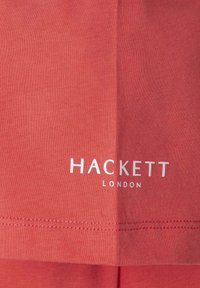 Korálová bavlnená tričko s hladkou textúrou, s bielym brandingom "HACKETT LONDON" na spodnej strane a jednoduchým, klasickým dizajnom.