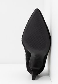 Suela de zapato de tacón de aguja negra con punta afilada, con agarre texturizado y marcas de la marca, inclinada contra una pared blanca en la esquina.