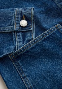 Tamsiai mėlynas džinsinio audinio paviršius su matoma tekstūra, auksinės siūlės, sidabrinio mygtuko užsegimas ir kišenės detalė viršuje kairėje.