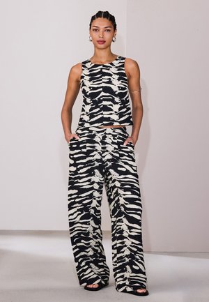 Žena s vrkočmi, ktorá nosí čierno-biele zebrované tielko bez rukávov a zodpovedajúce široké nohavice s rukami vo vreckách.