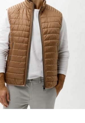 Mann trägt eine hellbraune gesteppte Weste über einem weißen Langarmshirt und hellgrauen Hosen, die Hände ruhen in der Nähe seiner Taschen.