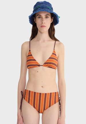 Kobieta ubrana w pomarańczowo-brązowy bikini w paski z wiązaniami po bokach i niebieski kapelusz typu bucket, stojąca na jednolitym jasnoszarym tle.