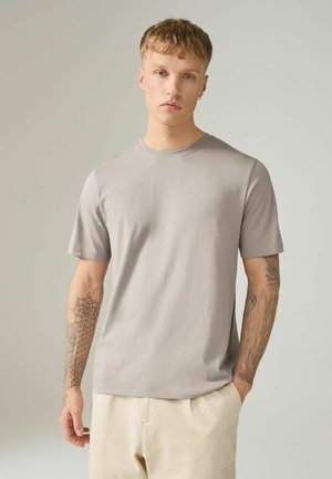 Bărbat tânăr cu păr scurt și tatuaje pe ambele brațe, purtând un tricou simplu gri deschis cu mâneci scurte și pantaloni bej pal.